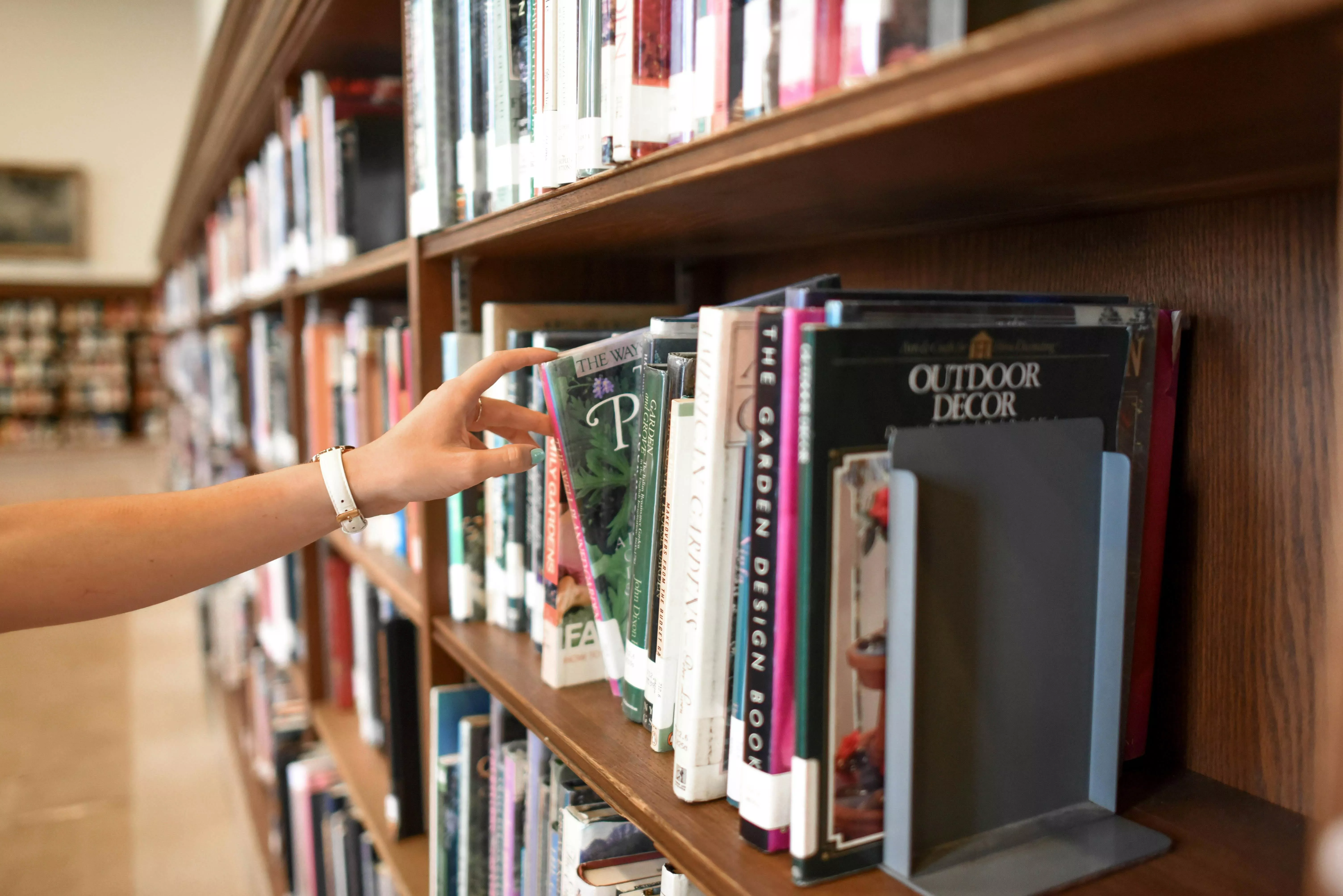 Photo of a hand reaching for a book from many books in the library stacks.